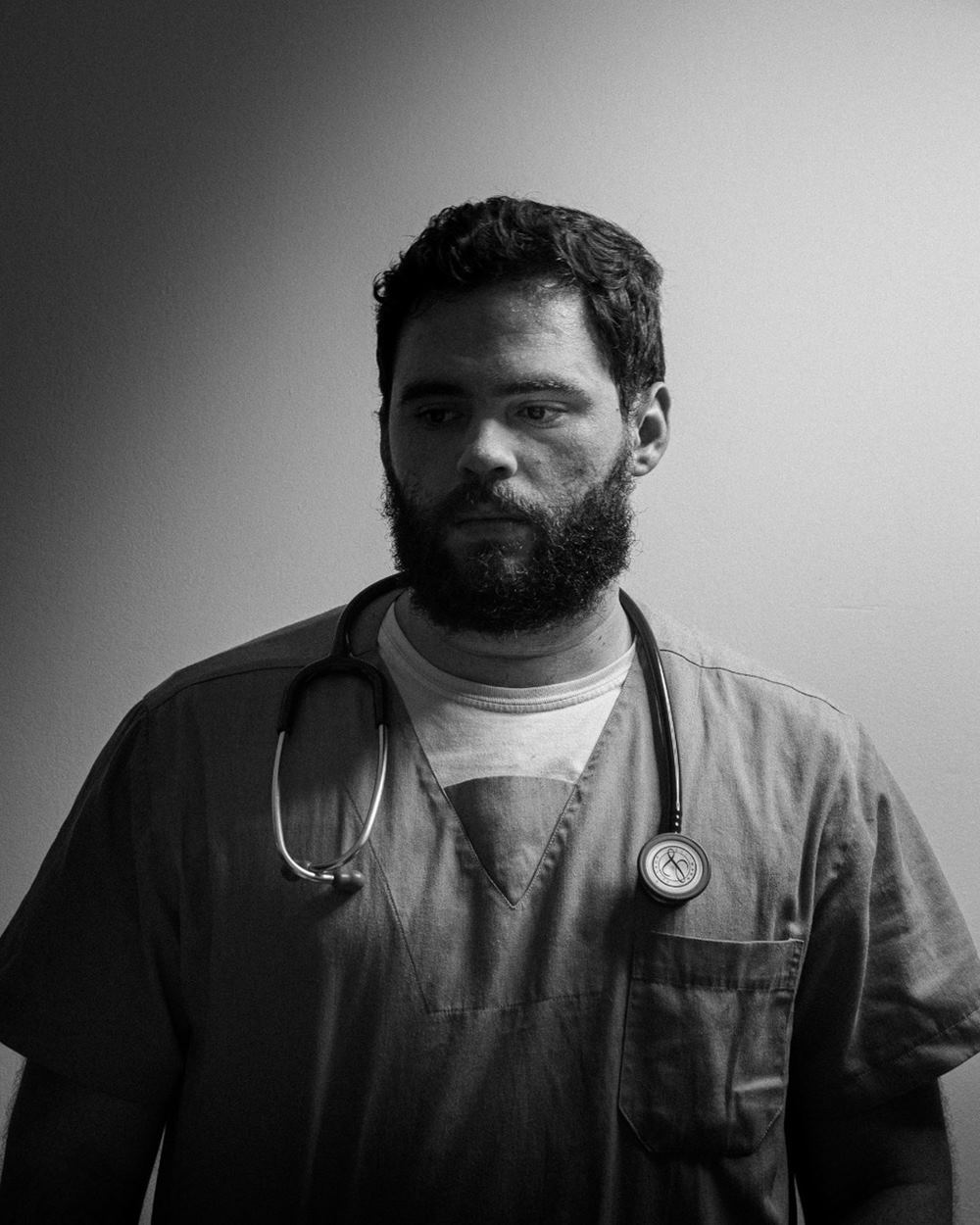 O influencer Vitor Arruda, um homem branco e jovem, de barba e cabelo preto, posa para foto em um fundo branco. Ele usa uma camiseta e um camisão, o tradicional uniforme de médico, e o instrumento estetoscópio no pescoço. - Metrópoles