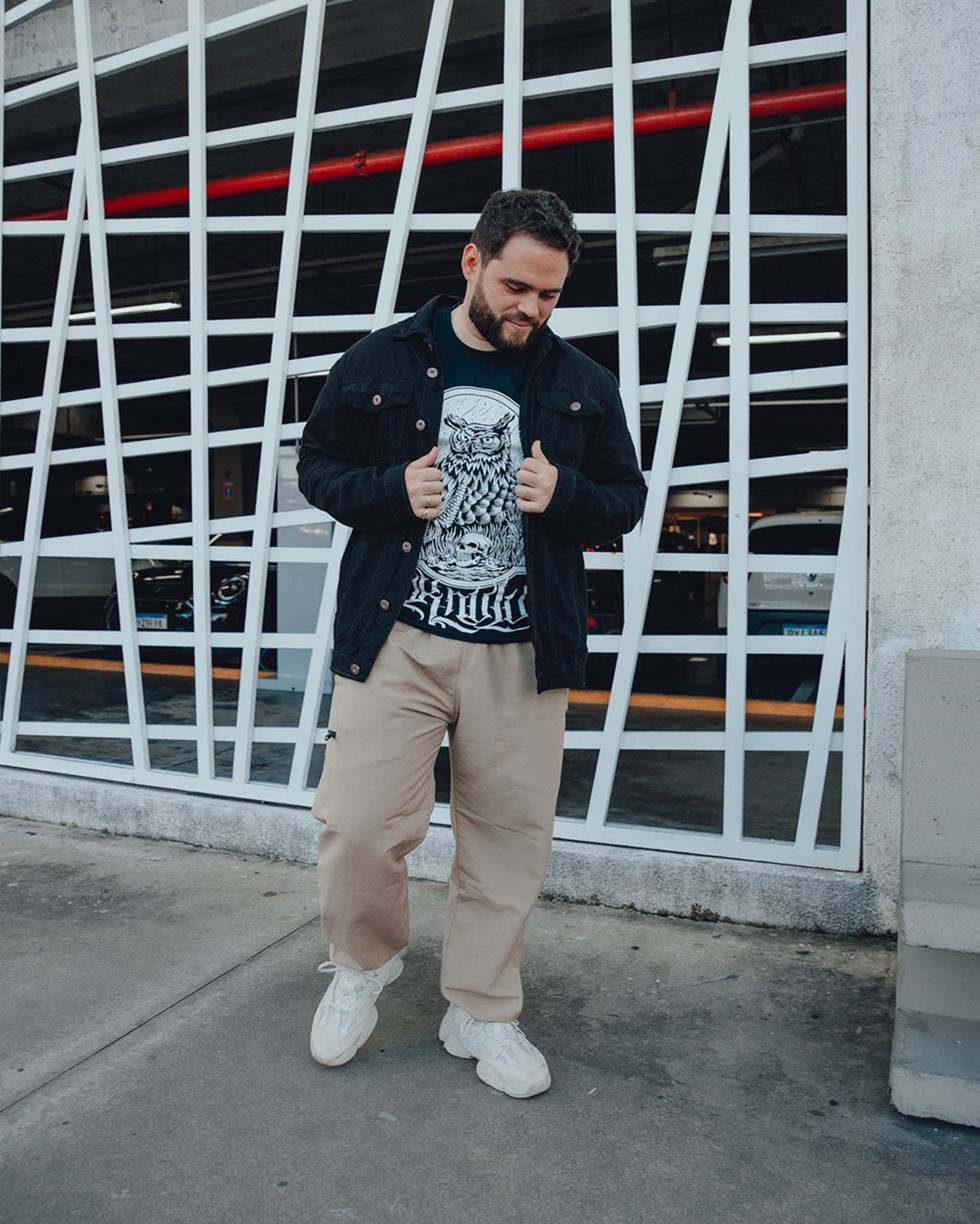 O influencer Vitor Arruda, um homem branco e jovem, de barba e cabelo preto, posa para foto em frente a um portão de ferro branco. Ele usa uma camiseta preta com uma coruja estampada; uma jaqueta jeans, também preta; calça bege; tênis branco Yeezy, do Kanye West e da marca Adidas. - Metrópoles