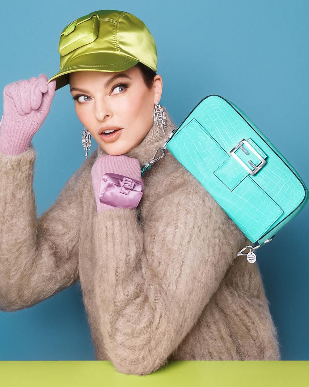 A modelo Linda Evangelista, uma mulher branca de meia idade, com olhos azuis e cabelos lisos, em foto de campanha da marca Fendi e Tiffany & Co. Ela segura uma bolsa modelo Baguette da marca Fendi feita em parceira com a Tiffany & Co. A peça é de couro e no tom de azul turquesa típico da joalheria. - Metrópoles