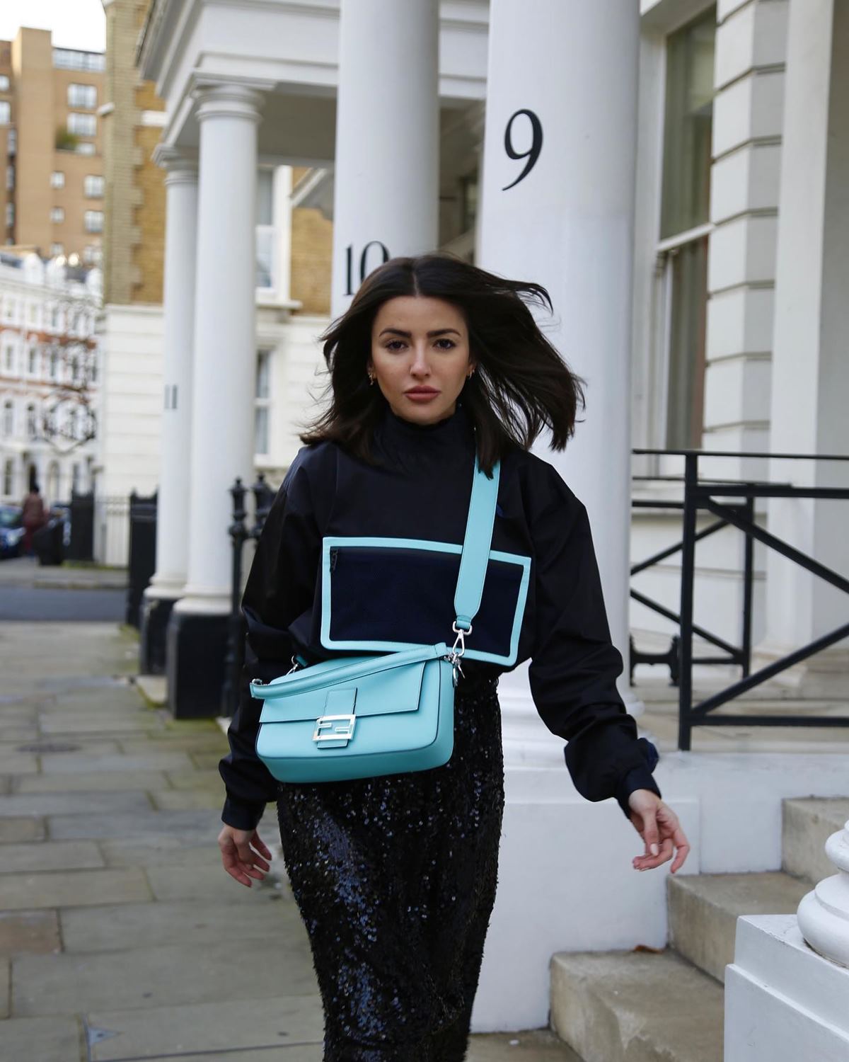 A influencer Alexandra Pereira, uma mulher branca jovem, de cabelo liso preto, posa para foto na rua. Ela usa casaco de moletom e calça, tudo preto, e uma bolsa modelo Baguette da marca Fendi feita em parceira com a Tiffany & Co. A peça é de couro e no tom de azul turquesa típico da joalheria. - Metrópoles