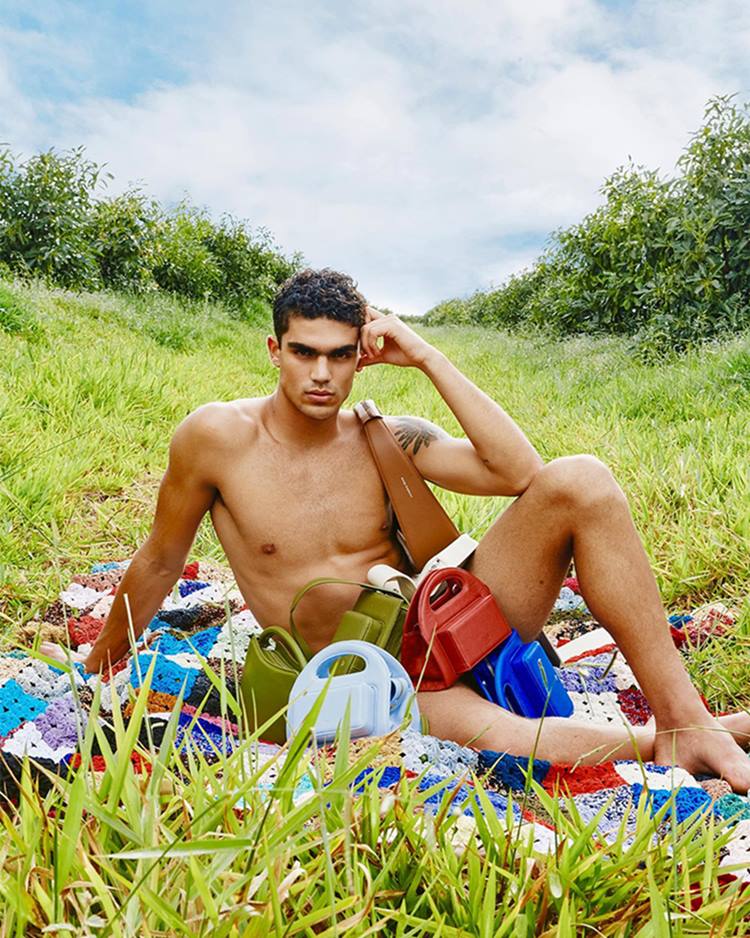 Homem moreno e jovem, de cabelo curto, posa para foto sentado em uma toalha no meio do jardim, simulando um picnic. Ele está semi nu e cobre a região da virilha com várias bolsas de couro coloridas da marca Misci. - Metrópoles