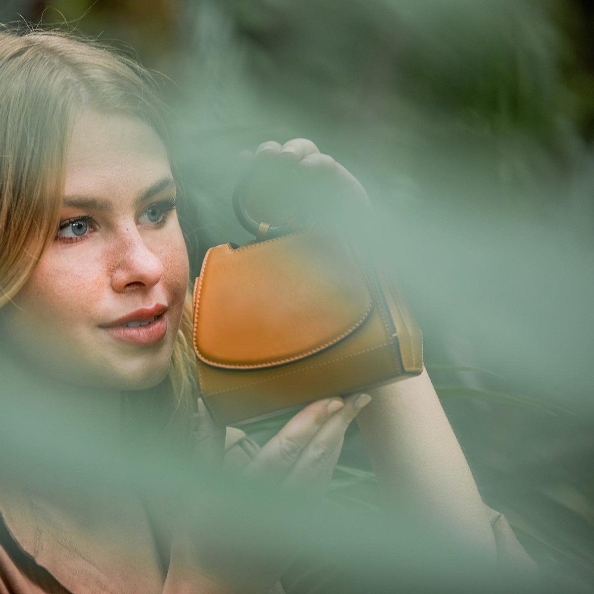 Mulher branca e jovem, de cabelo liso loiro, com olhos azuis. Ela posa para foto e um jardim. Segura uma bolsa de couro marrom, com alça de madeira, em tamanho mini. - Metrópoles