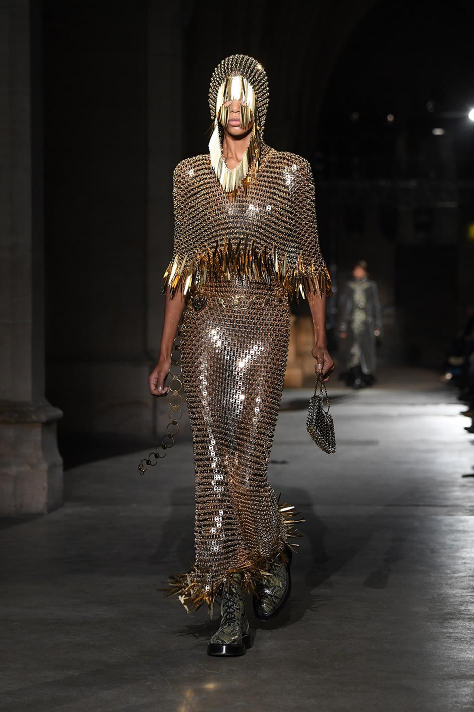 Desfile de outono/inverno 2020/2021 da marca Paco Rabanne. Na imagem, uma modelo negra anda pela passarela com um vestido longo e um adereço que cobre o rosto. As duas peças são douradas. - Metrópoles