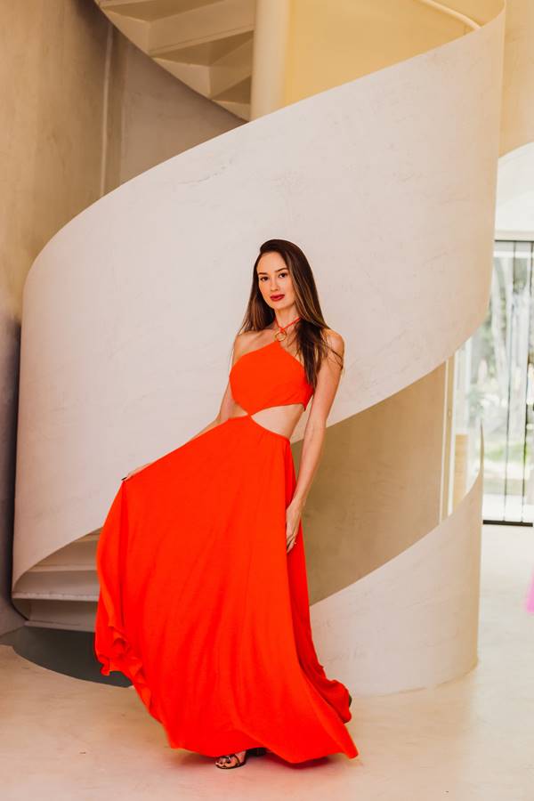 Cecília Rosa encostada na escada usa vestido laranja com recortes na altura das costelas, cabelos para trás. O vestido está em movimento - Metrópoles