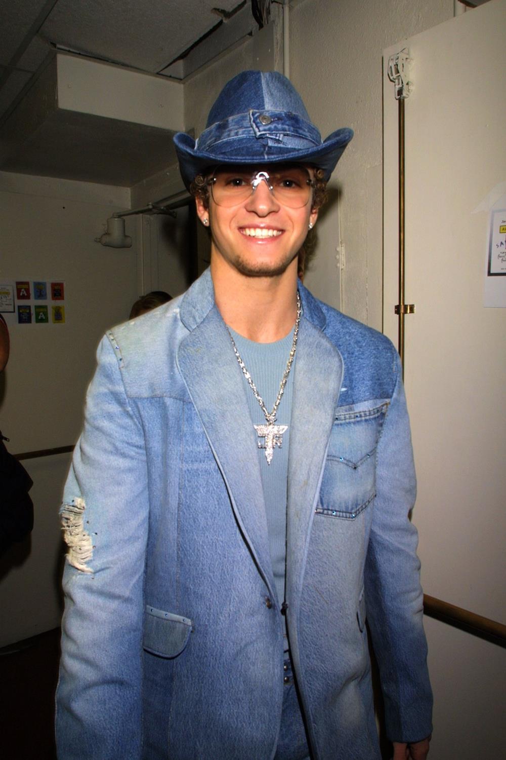 O cantor Justin Timberlake na premiação Video Music Awards (VMA) de 2001. Ele é branca, jovem e loiro e usa um chapéu, uma blusa e um blazer. Todas as peças são no tecido jeans. Usa ainda um óculos escuros com lente marrom transparente. - Metrópoles