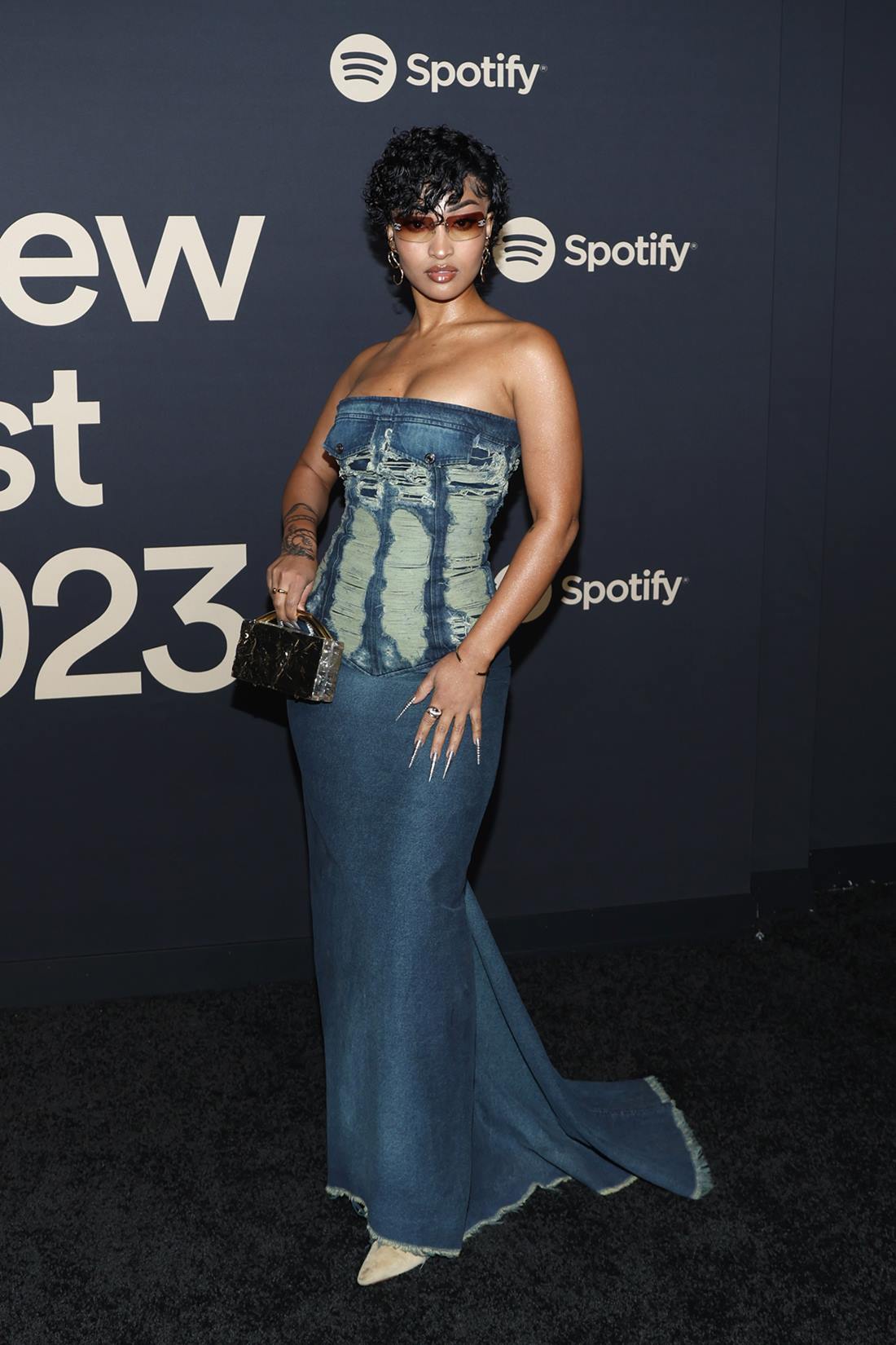 A cantora Shenseea, uma mulher negra e jovem, de cabelo preto curto e cacheado, na festa do Spotify. Ela usa um vestido longo jeans, em lavagem escura, uma bolsa preta de couro e óculos escuros. - Metrópoles