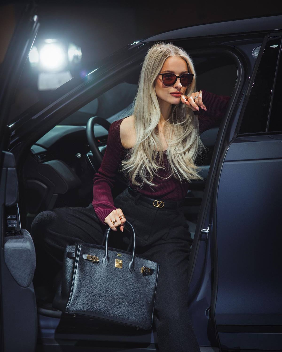 Mulher branca e jovem, de cabelo liso loiro, posa para foto sentada no banco de um carro. Ela usa uma blusa de manga longa vinho, uma calça preta, um óculos escuro e uma bolsa preta da marca Hermès. - Metrópoles