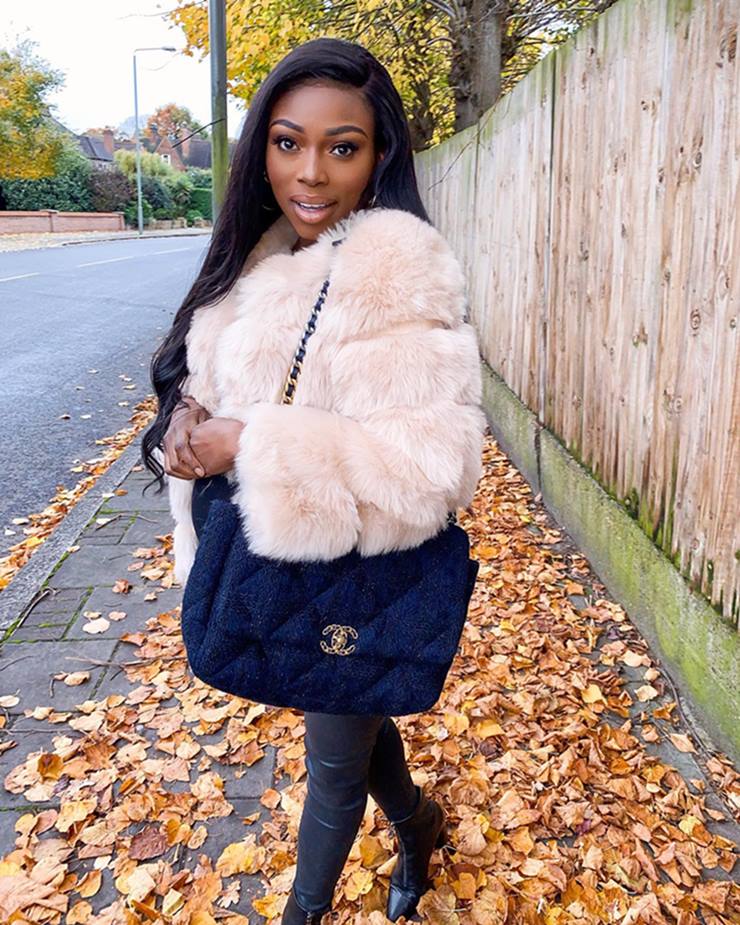 Mulher negra e jovem, de cabelo liso preto, posa para foto nas ruas de Londres. Ela usa umaum casaco de pelo bege, uma calça preta de couro e uma bolsa de pano da marca Chanel. - Metrópoles