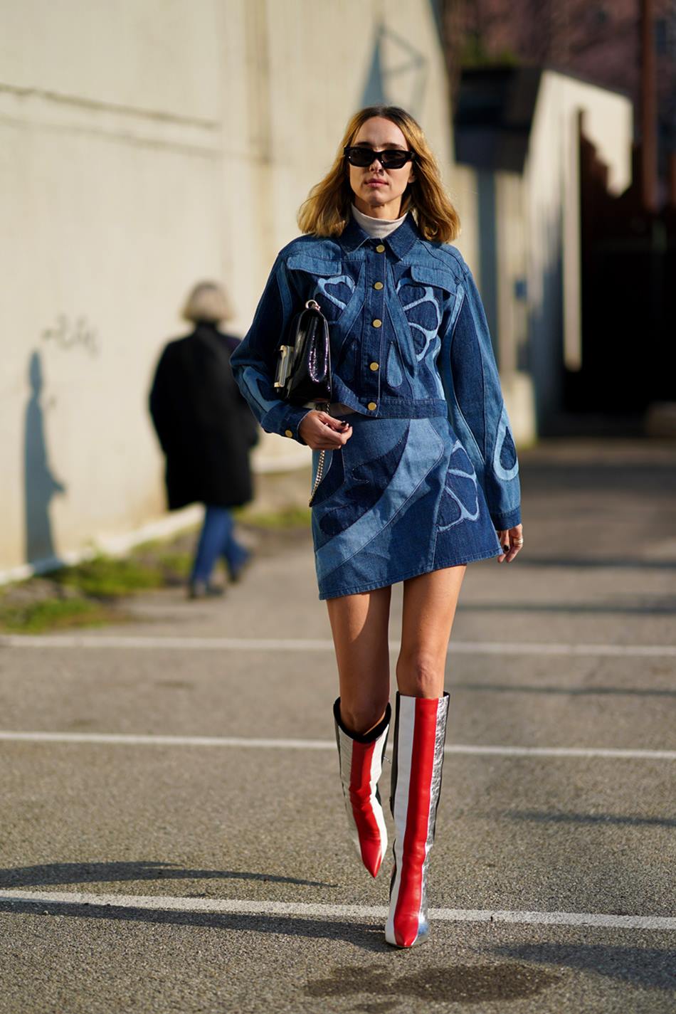 Mulher branca e jovem, de cabelo liso loiro, posa para foto andando nas ruas durante a Semana de Moda de Milão. Ela usa um conjunto de jaqueta e saia, ambos em jeans, com uma estampa floral; óculos escuros; bota de couro e uma bolsa preta, também de couro. - Metrópoles