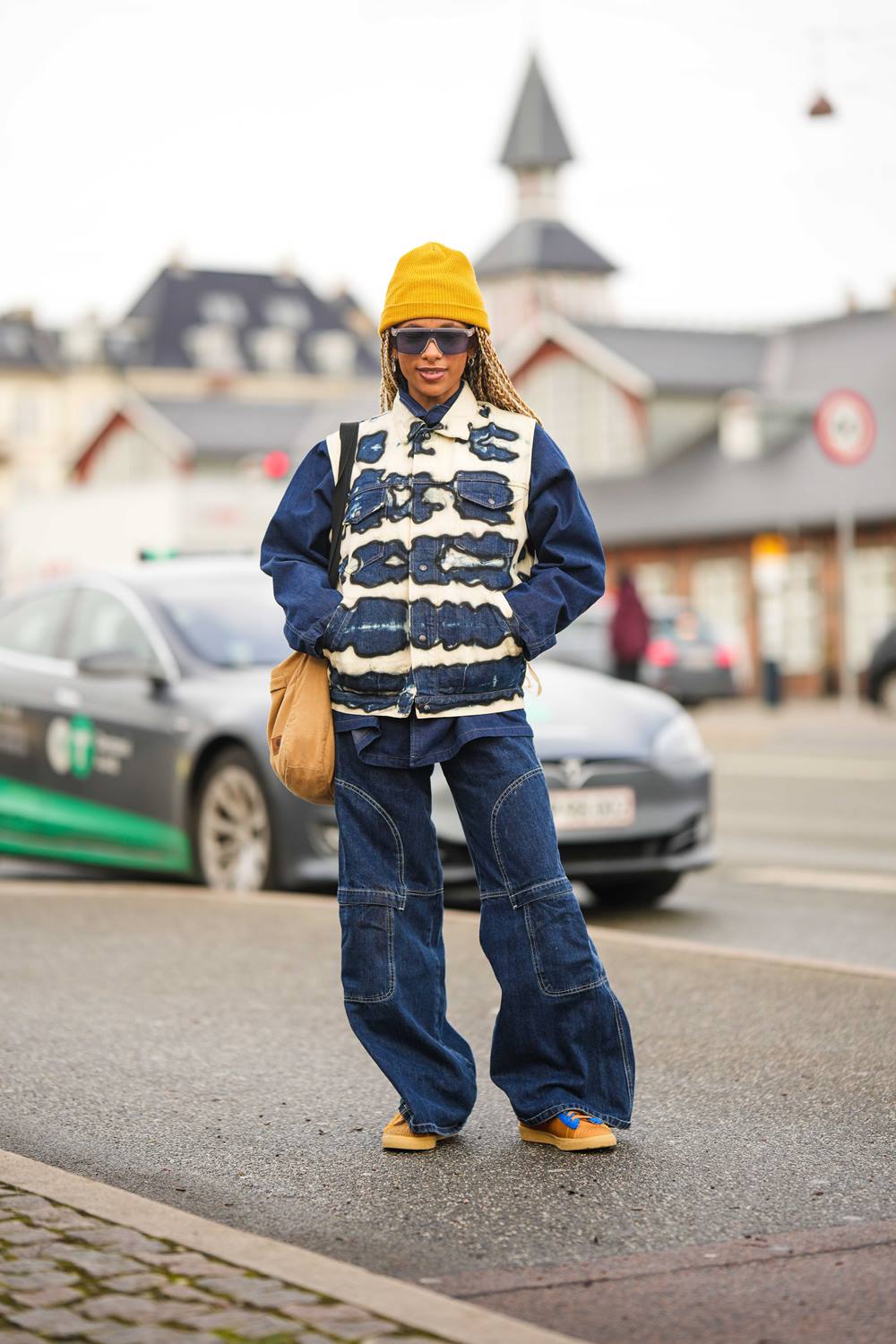 Mulher jovem e negra, de cabelos trançados longos e loiros, posa para foto em frente a casas. Ela usa uma jaqueta jeans, com detalhes descoloridos em branco; uma calça jeans; óculos escuros; uma touca de tricô amarelo e um tênis caramelo de camurça. - Metrópoles