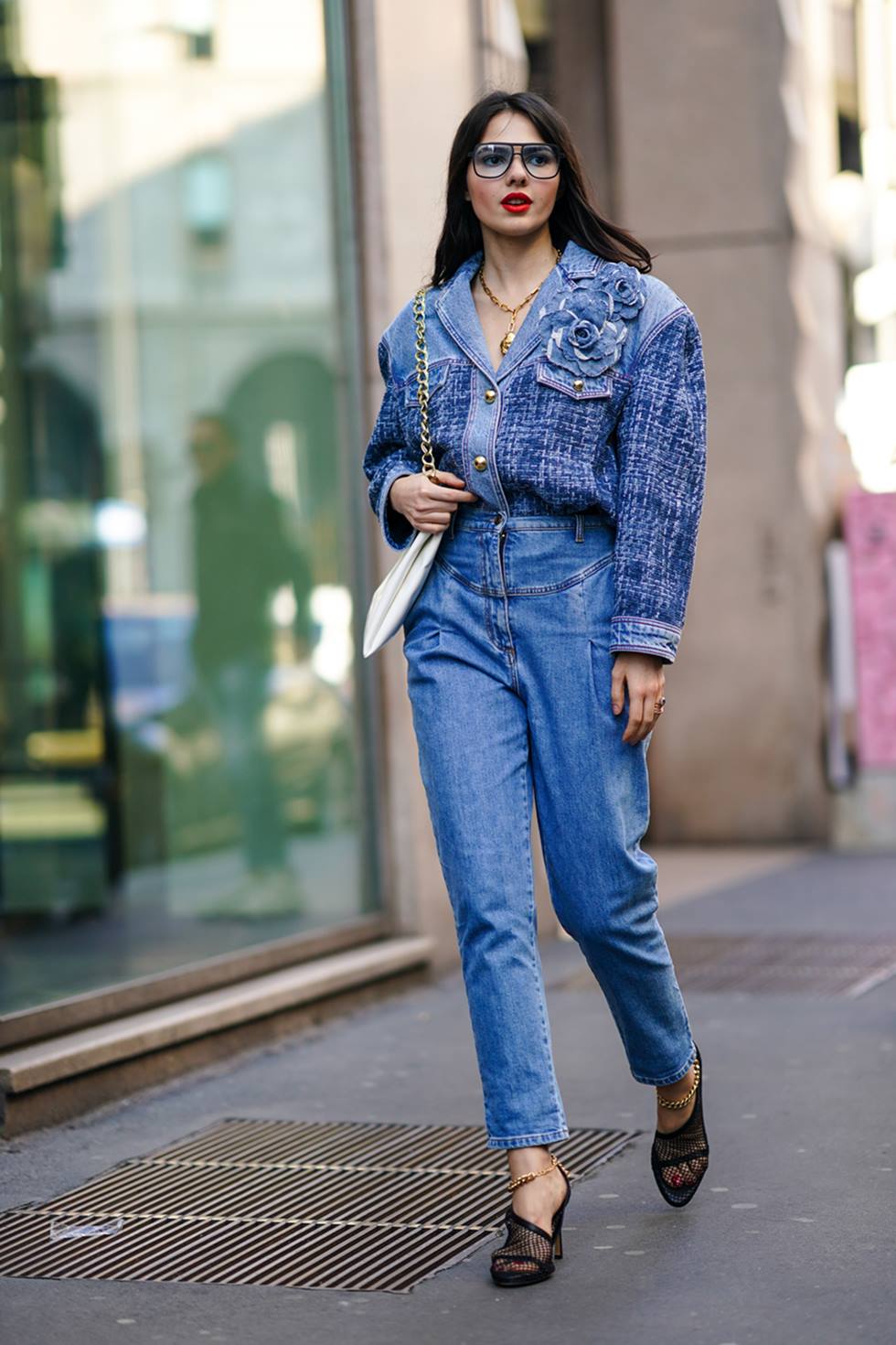 Mulher branca e jovem, de cabelo liso castanho, anda pela rua durante a Semana de Moda de Milão. Ela usa uma jaqueta e uma calça, ambas em jeans; uma sandálai de salto preta; uma bolsa de couro branca e um óculos de grau com armação preta. - Metrópoles