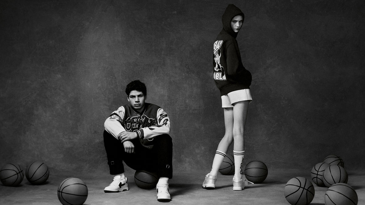 Campanha de divulgação da coleção de roupas da marca Off-White com o time de basquete Chicago Bulls. Na imagem, é possível ver uma mulher e um homem, ambos brancos jovens, posando para foto em um fundo cinza com bolas de basquete. Ambos usam tênis esportivos e casacos moletons preto com o desenho do mascote do time de basquete. - Metrópoles