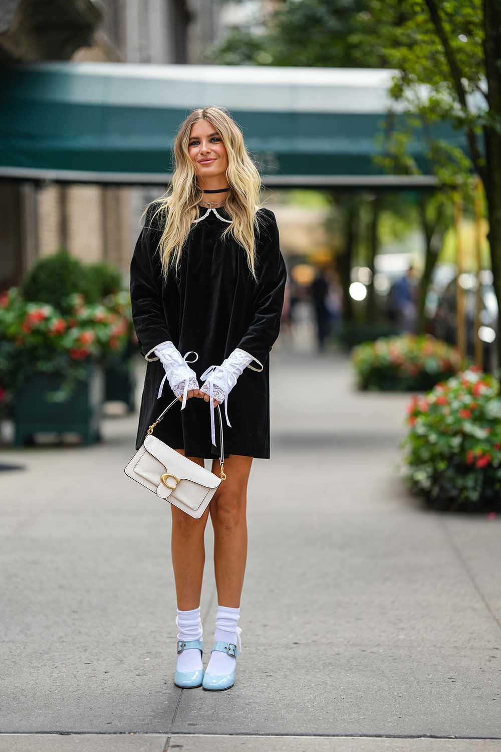 Mulher branca e jovem, de cabelo liso loiro e longo, posa para foto nas ruas de Nova York. Ela usa um vestido preto de camurça, uma meia branca, uma sandália de salto azul clara e uma bolsa branca, de couro, do modelo Tabby da marca Coach. - Metrópoles