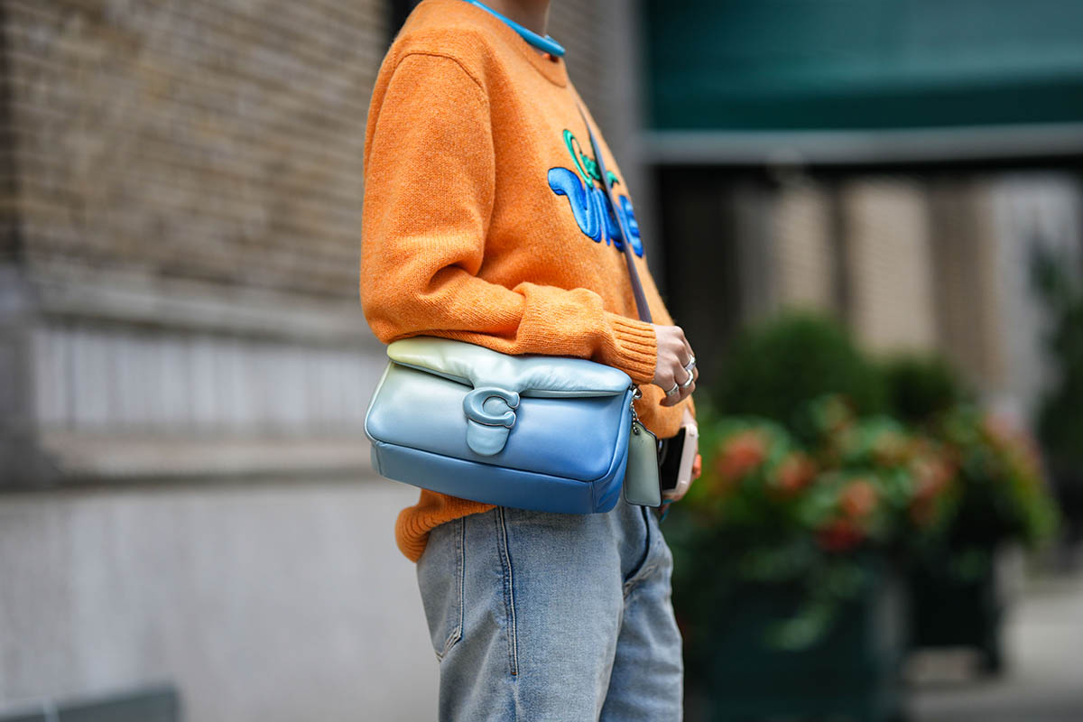 Pessoa posa para foto durante a Semana de Moda de Nova York. Não é possível ver seu rosto, apenas suas mãos, que são brancas. Usa um casaco de tricô laranja com detalhes azuis, calça jeans e uma bolsa de couro azul em degradê, modelo da Tabby, da marca Coach. - Metrópoles