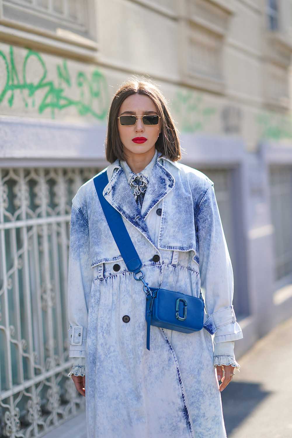 Mulher branca e jovem, de cabelo preto liso, posa para foto na rua. Ela usa um vestido longo, feito em jeans; óculos escuros; bolsa de couro azul, modelo Snapshot, da marca Marc Jacobs. - Metrópoles