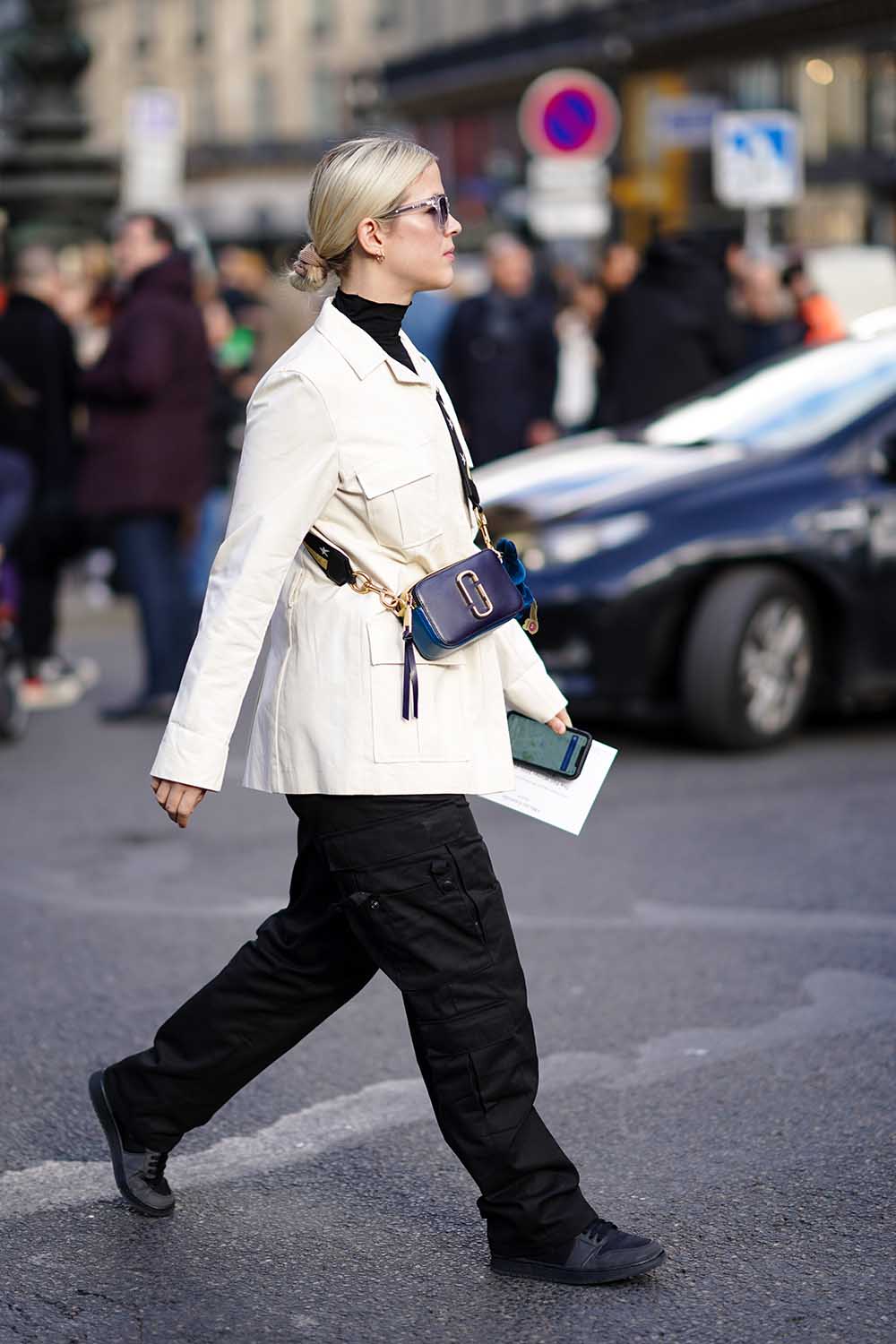 Mulher branca e loira, de cabelo liso loiro amarrado, anda pelas ruas durante a Semana de Moda de Paris. Usa uma calça preta; um tênis preto; blazer branco; óculos escuros; e uma bolsa modelo Snapshot, em couro branco, da marca Marc Jacobs. - Metrópoles