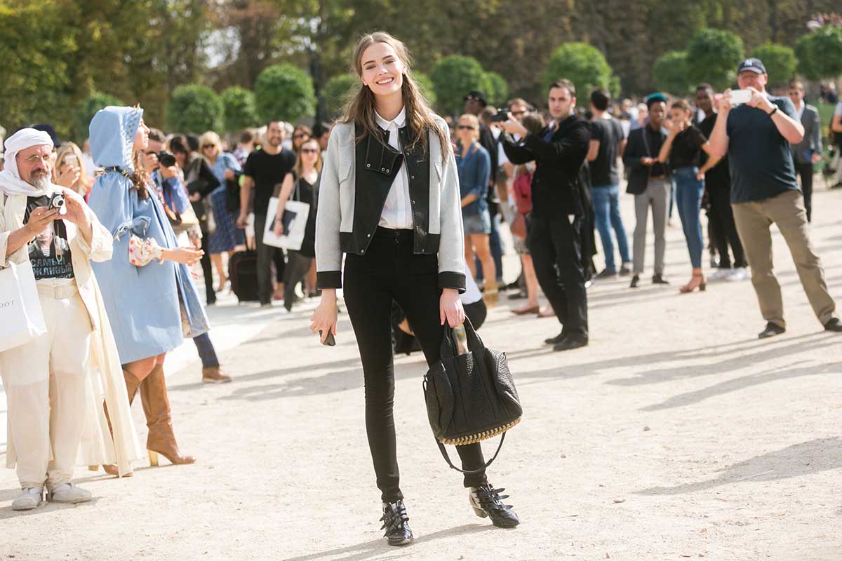 Mulher branca e jovem, de cabelo liso longo e castanho, posa para foto na rua durante a Semana de Moda de Paris. Usa uma camisa branca, calça preta, bota preta de couro com fivelas, uma jaqueta de couro preta com cinza e a bolsa Rocco, em couro preto, da marca Alexander Wang. - Metrópoles