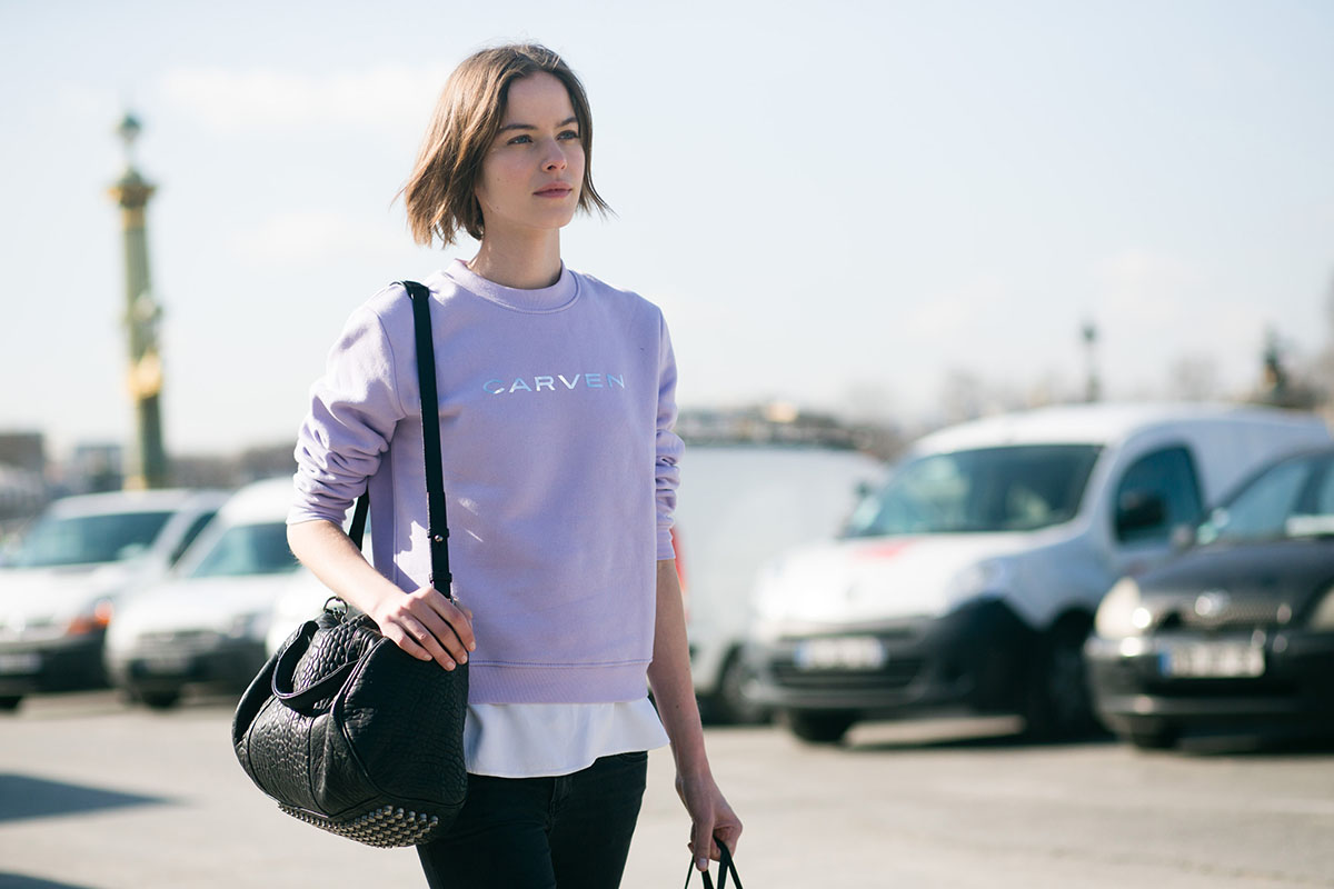 Mulher branca e jovem, de cabelo liso curto e castanho, anda por uma rua. Ela usa um moletom lilás, calça jeans preta e segura uma bolsa preta, de couro, da marca Alexander Wang. O modelo se chama Rocco. - Metrópoles