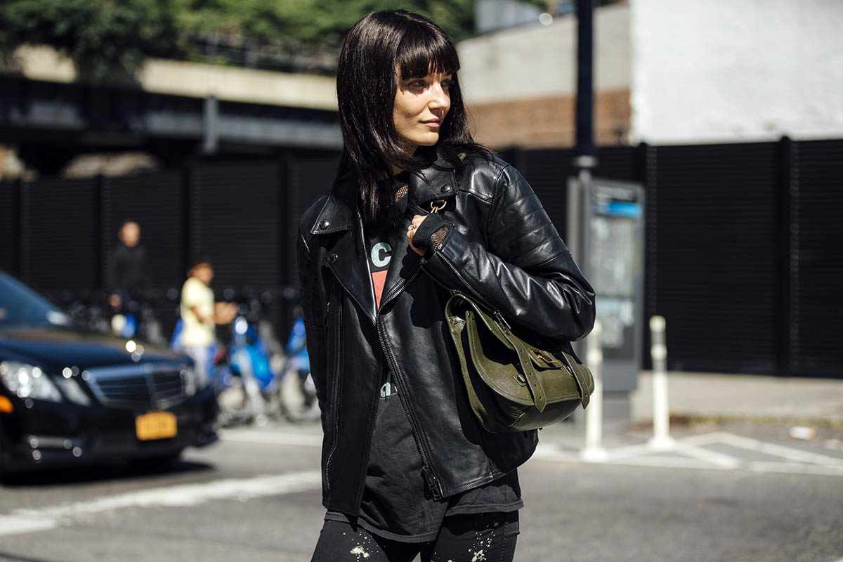 Mulher branca e jovem, de cabelo preto liso com franja, andando pela rua. Ela usa uma camiseta, uma calça jeans e uma jaqueta de couro; todas as peças são na cor preta. Segura uma bolsa verde musgo em couro, modelo PS1, da marca Proenza Schouler. - Metrópoles