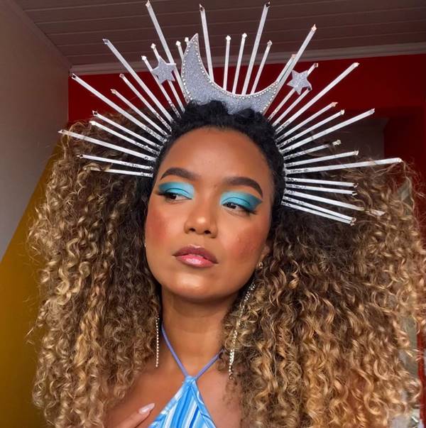 Mulher negra usa tirara com lua e linhas pontudas, sombra azul nos olhos e blusa frente única azul. Ela tem cabelos com mechas loiras e cachos - Metrópoles 