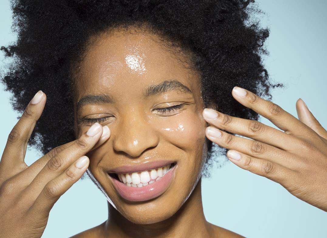 Mulher negra de olhos fechados enquanto passa um produto de textura aquosa no rosto com as duas mãos