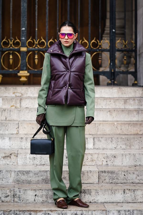 No street style, mulher usa conjunto de alfaiataria verde com blazer e calça. Por cima, um colete puffer bordô. Ela segura uma bolsa preta e usa óculos escuros com lentes amareladas - Metrópoles