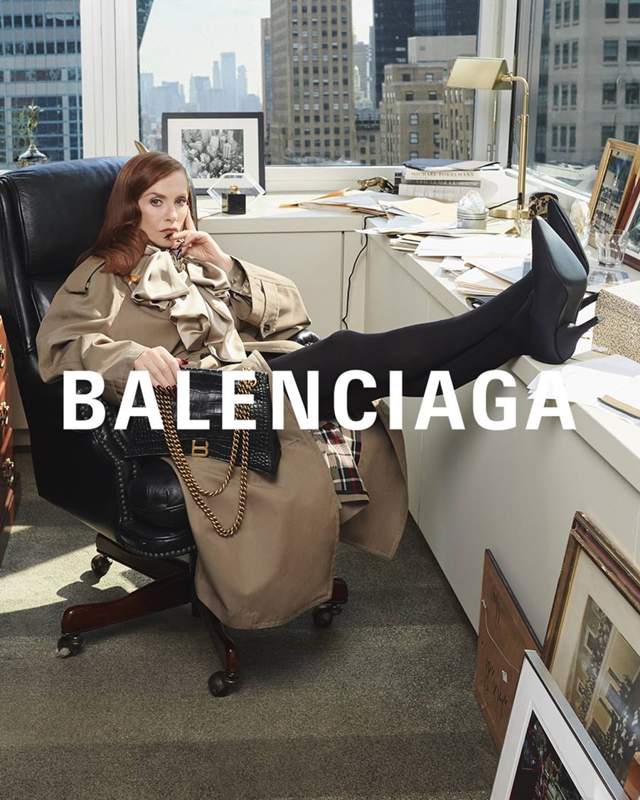 Campanha publicitária da Balenciaga. Na imagem, uma mulher branca e jovem, de cabelo ruivo, sentada em uma cadeira de escritório com os pés apoiados na mesa. Ela usa um casaco marrom, calça preta e bolsa preta de salto fino. - Metrópoles