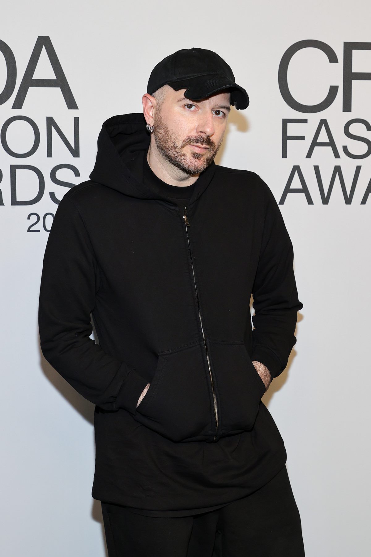 O estilista Demna Gvasalia, um homem jovem e branco, com cabelo raspado e barba castanha, em um evento de moda de 2021. Ele usa uma camiseta, uma calça jeans, um casaco moletom com capuz e um boné. Todas as peças são pretas. - Metrópoles