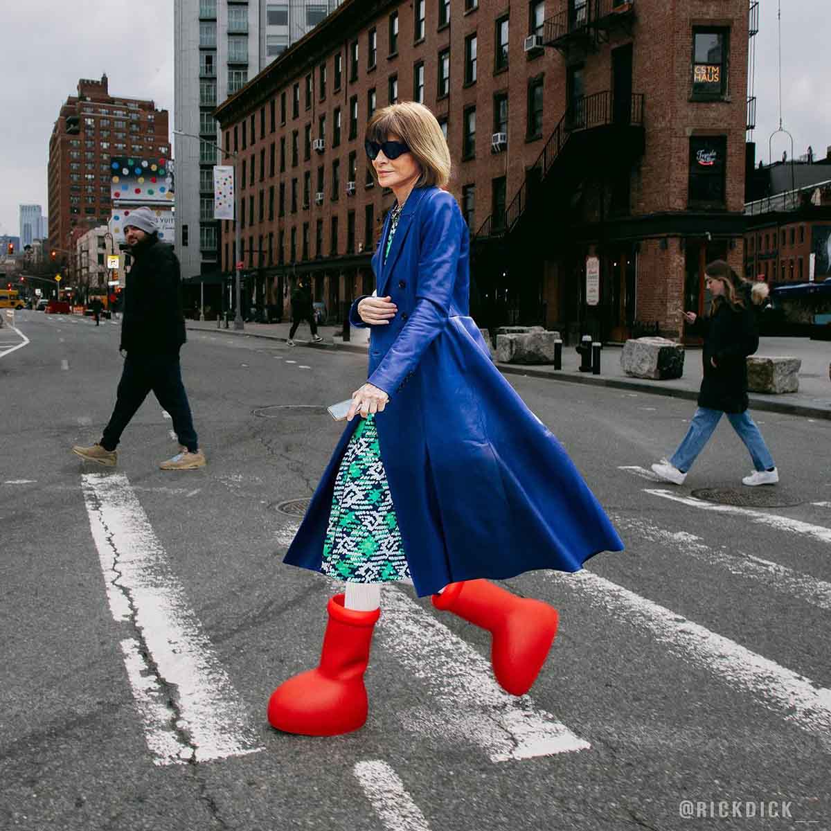 A jornalista de moda Anna Wintour, uma mulher branca, idosa e com cabelo liso loiro, andando pelas ruas de Nova York. Ela usa um vestido verde, um casaco azul e a bota vermelha gigante de plástico da marca MSCHF. - Metrópoles