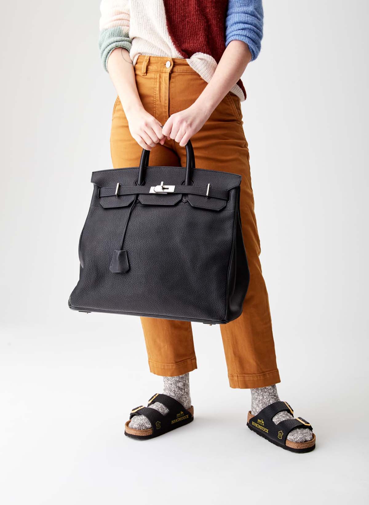 Campanha de divulgação da sandália Birkinstock, da marca MSCHF. O calçado é uma sandália baixa, feita de cortiça e couro. Na foto, uma pessoa usa a peça e veste uma calça marrom e segura a bolsa preta de couro Birkin, da grife Hermés. - Metrópoles