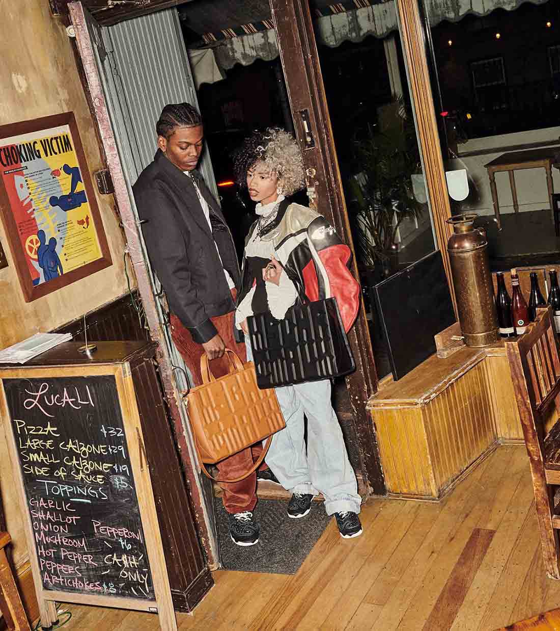 Campanha de divulgação da coleção Made In Italy, da marca MSCHF. Na foto, um homem e uma mulher, ambos negros, estão em um restaurante. Eles seguram bolsas de couro da marca, uma em preto e outra caramelo. - Metrópoles