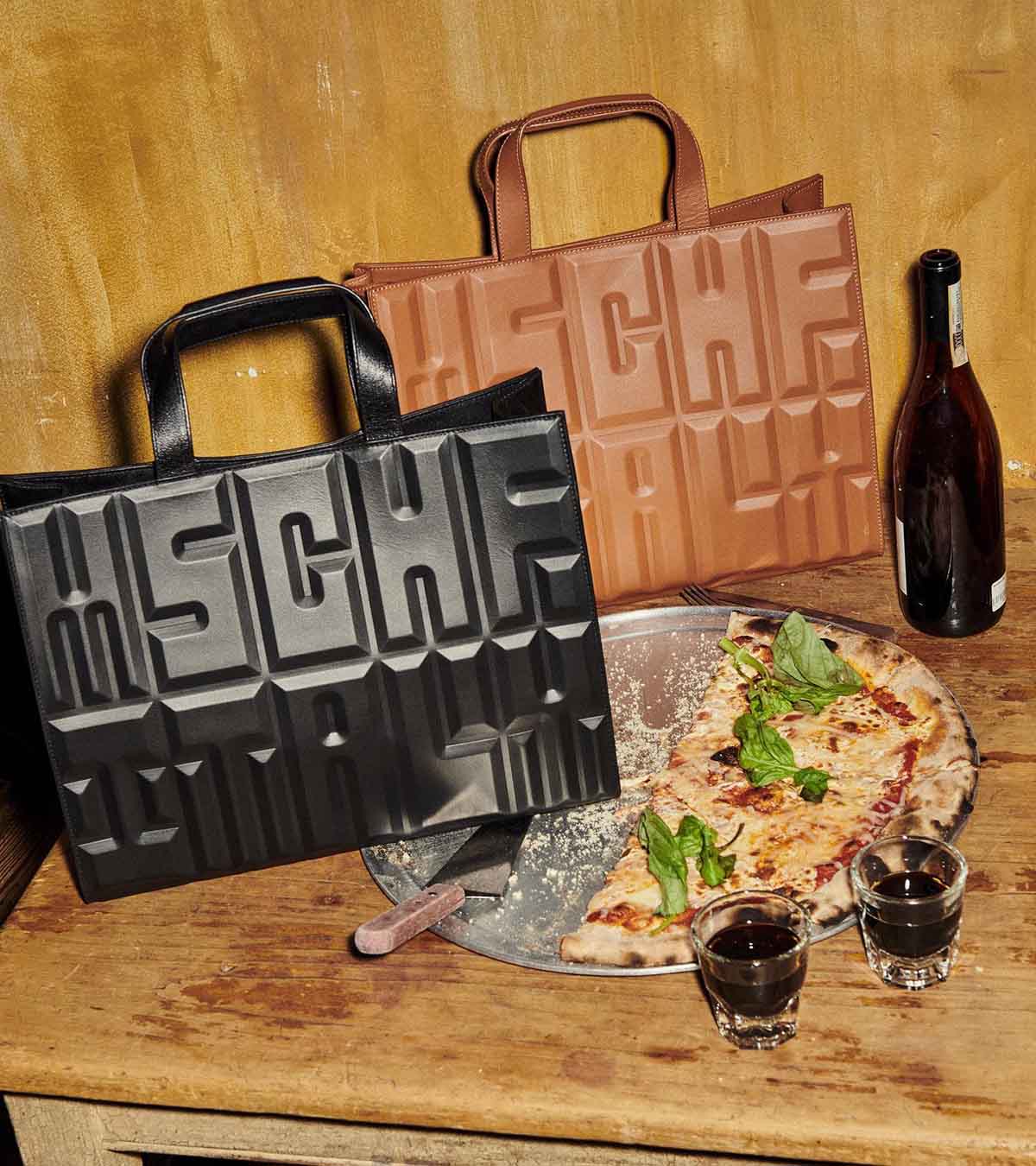 Campanha de divulgação da coleção Made In Italy, da marca MSCHF. Na foto, duas bolsas de couro da marca, uma em preto e outra caramelo, estão na mesa de um restaurante. É possível ver ainda duas taças de vinho e uma forma com metade de uma pizza. - Metrópoles