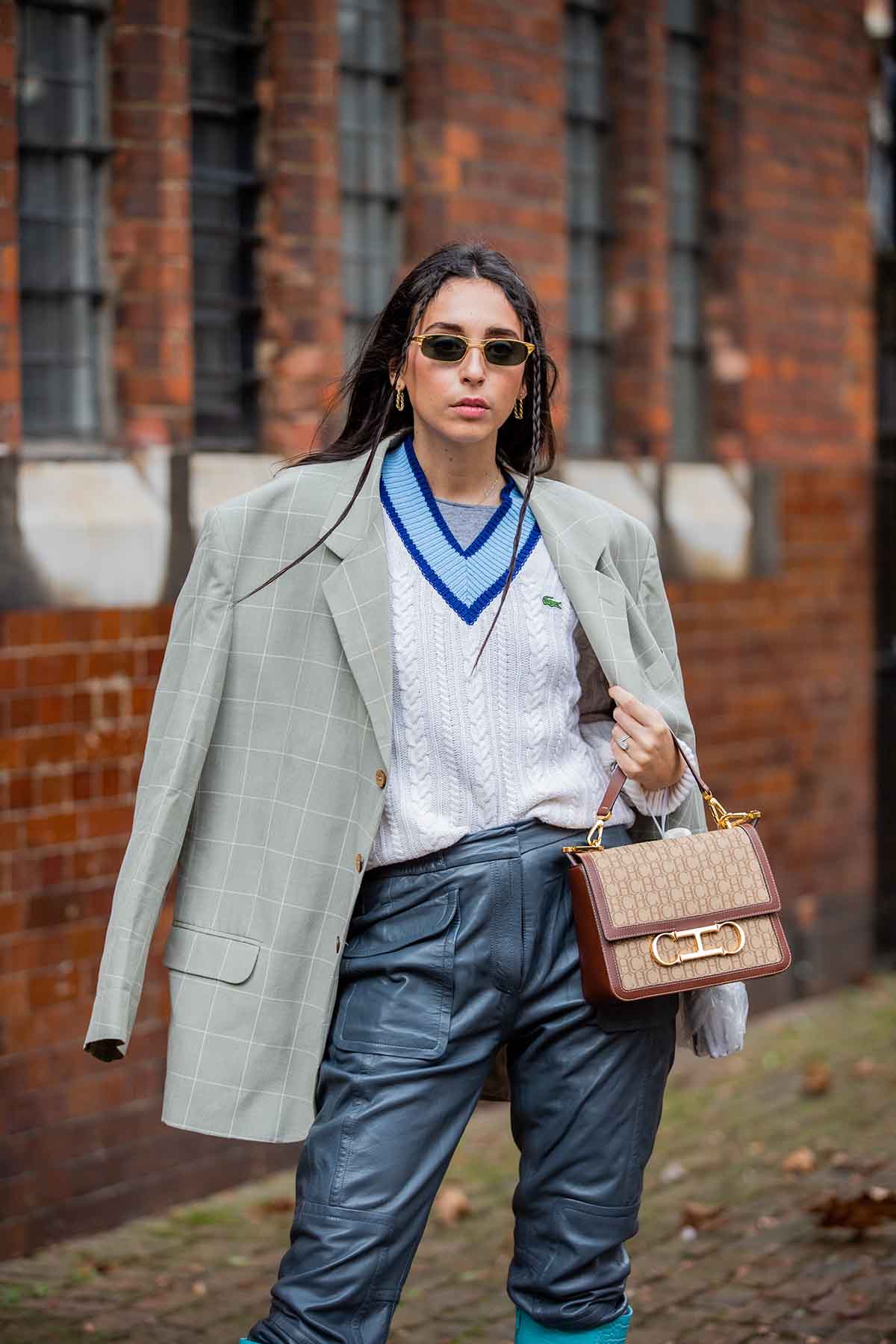 Mulher branca e jovem, de cabelo liso longo, castanho, posa para foto nas r uas de Londres. Ela usa um colete de tricô da marca Lacoste, calça preta de couro, blazer cinza e uma bolsa marrom de couro. - Metrópoles