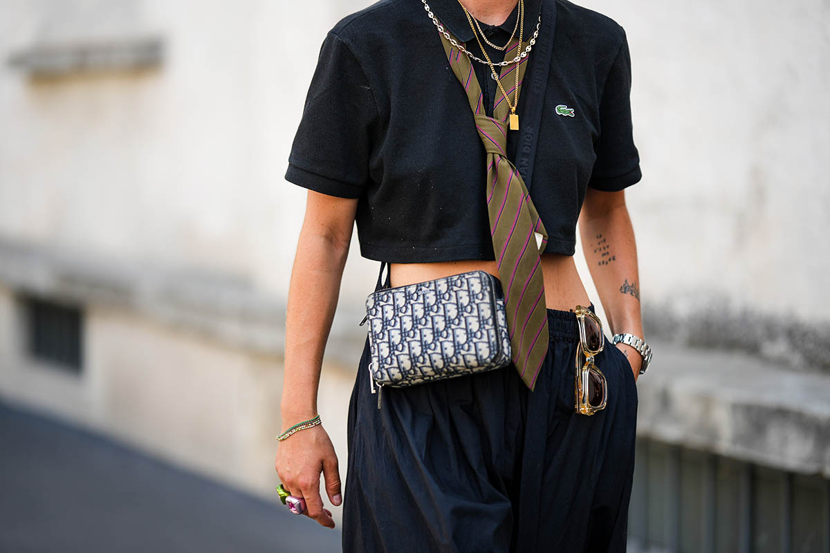 Mulher jovem e branca andando pela rua. Na imagem não é possível ver seu rosto, apenas do seu pescoço para o joelho. Ela usa uma camisa cropped preta da Lacoste, calça preta, uma gravata verde musgo e uma bolsa transpassada da Dior. - Metrópoles