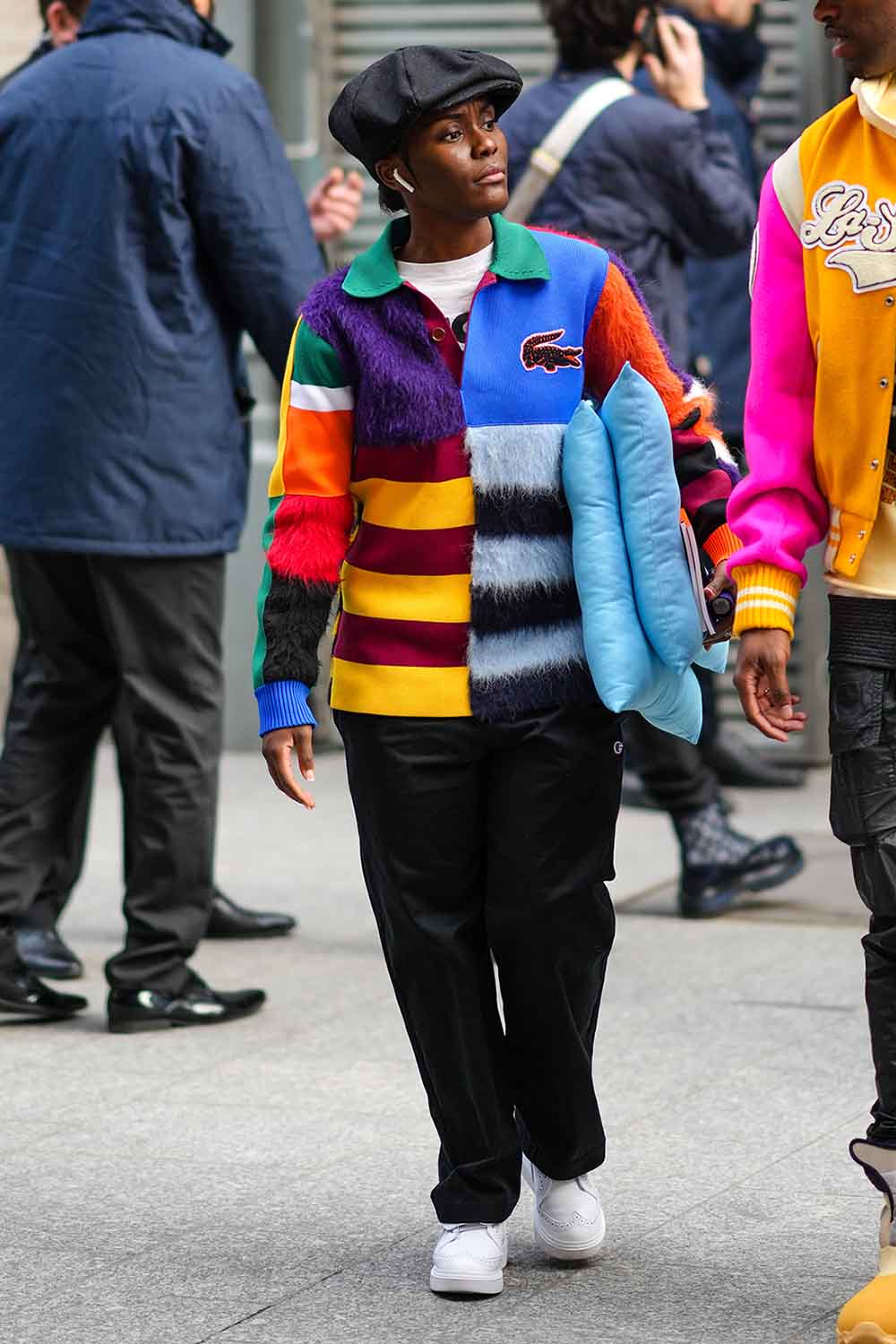 Mulher negra e jovem chegando no desfile da marca Louis Vuitton, na Semana de Moda de Paris. Ela usa uma calça preta, uma boina, também preto, um tênis branco de couro e u mcasaco estampado da marca Lacoste. - Metrópoles