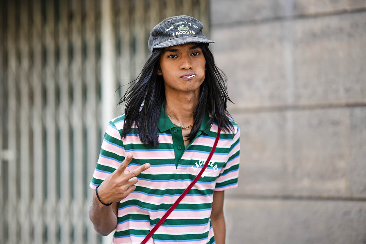 Homem jovem e negro, de cabelos longos e lisos, andando pela rua. Ele usa uma camisa polo listrada, nas cores verde, rosa e branco; e um boné cinza. Todas as peças são da marca Lacoste, com o logo do jacaré. - Metrópoles