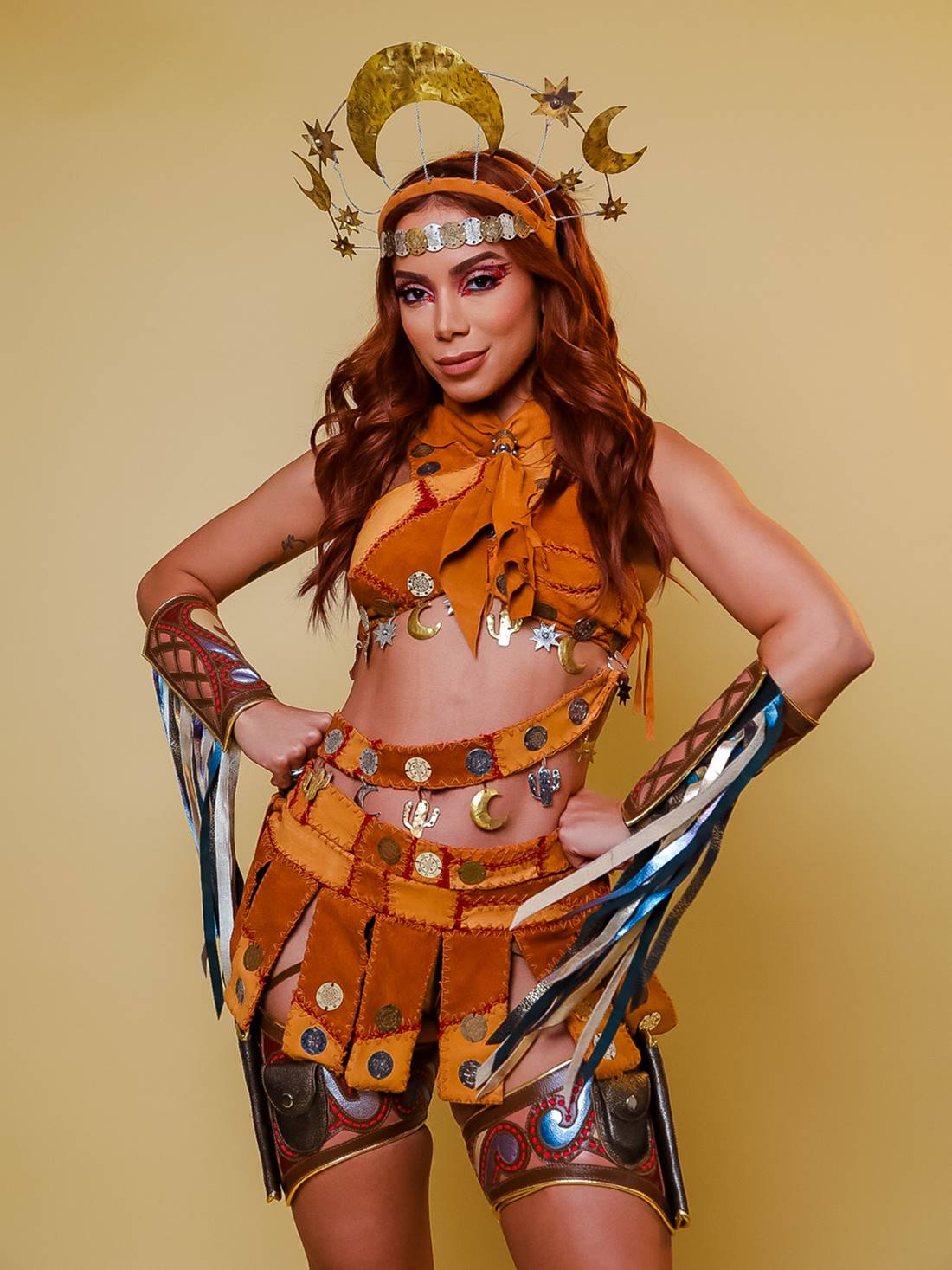 A cantora Anitta, uma mulher parda e jovem, de cabelo ondulado longo e acobreado, posa para foto em um fundo bege liso. Ela usa um top e uma saia de couro, além de uma tiara tourada. A roupa faz uma referência ao uniforme do cangaço usado por Maria Bonita. - Metrópoles