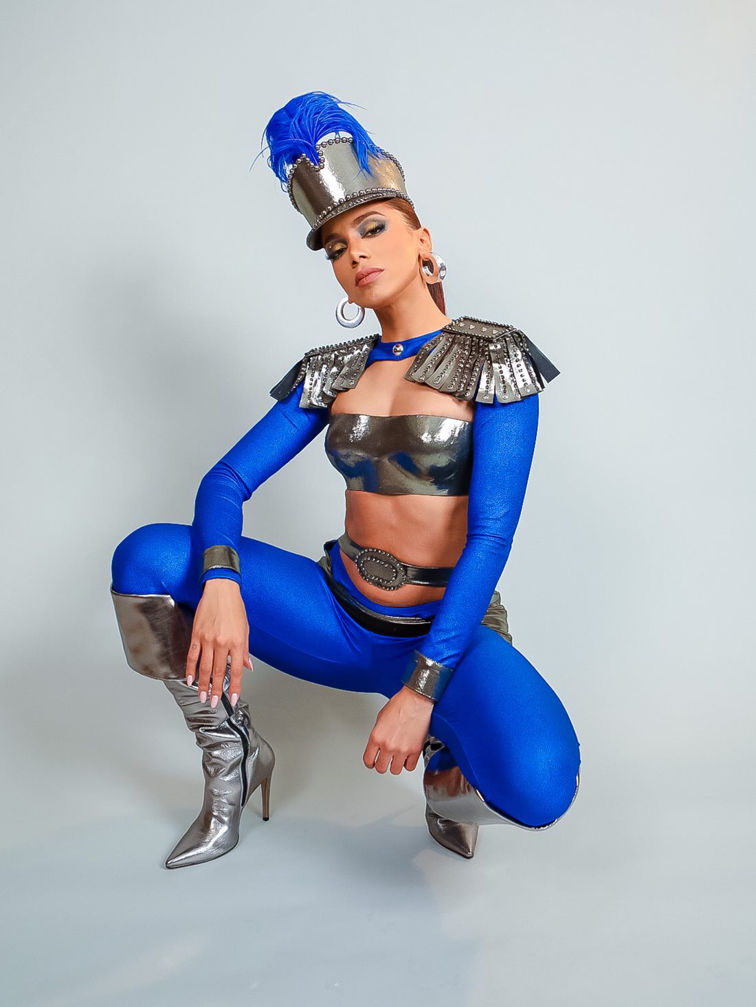 A cantora Anitta, uma mulher parda e jovem, de cabelo ondulado longo e acobreado, posa para foto em um fundo cinza liso. Ela usa um top prata e uma legging azul, além um chapéu que lembra o usado por militares no século XIX. A roupa faz uma referência à guerreira Maria Quitéria. - Metrópoles