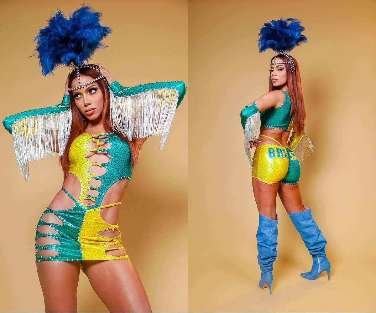 A cantora Anitta, uma mulher parda e jovem, de cabelo ondulado longo e acobreado, posa para foto em um fundo bege liso. Ela usa um vestido verde e amarelo, com decotes no centro, uma luva azul e um acessório de Carnaval azul com penas na cabeça. - Metrópoles