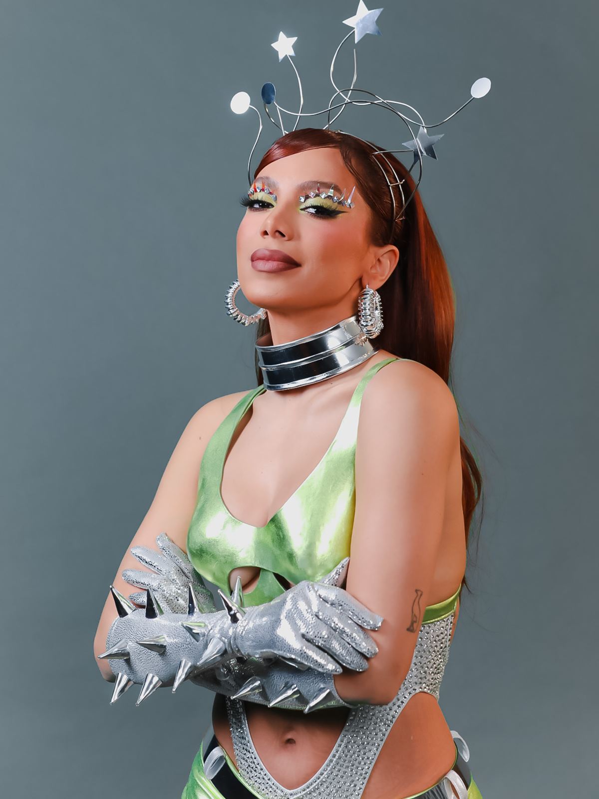 A cantora Anitta, uma mulher parda e jovem, de cabelo ondulado longo e acobreado, posa para foto em um fundo bege liso. Ela usa um top e uma saia, ambos verde metalizado, e botas grandes na mesma cor; além de uma tiara prateada. - Metrópoles