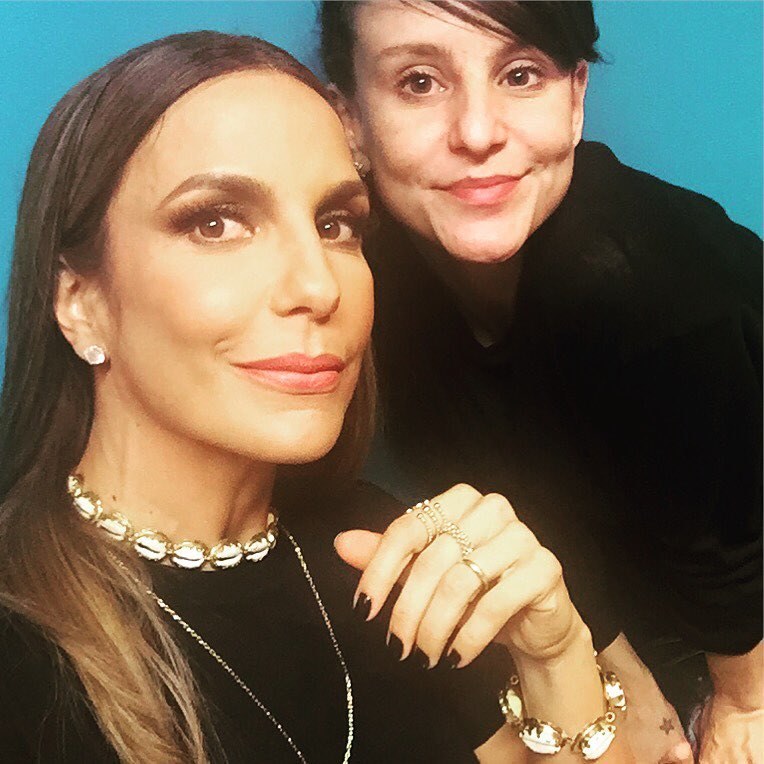 A stylist Clara Lima, uma mulher branca e jovem, de cabelos ondulados castanhos e franja, ao lado de Ivete Sangalo. A cantora baiana é uma mulher morena, de meia idade, com cabelo liso longo. Ambas usam roupas pretas. - Metrópoles