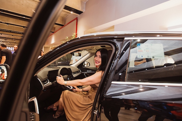 Mulher posando para foto dentro de um carro parado, com a porta do motorista aberta e sorrindo com as mãos ao volante 