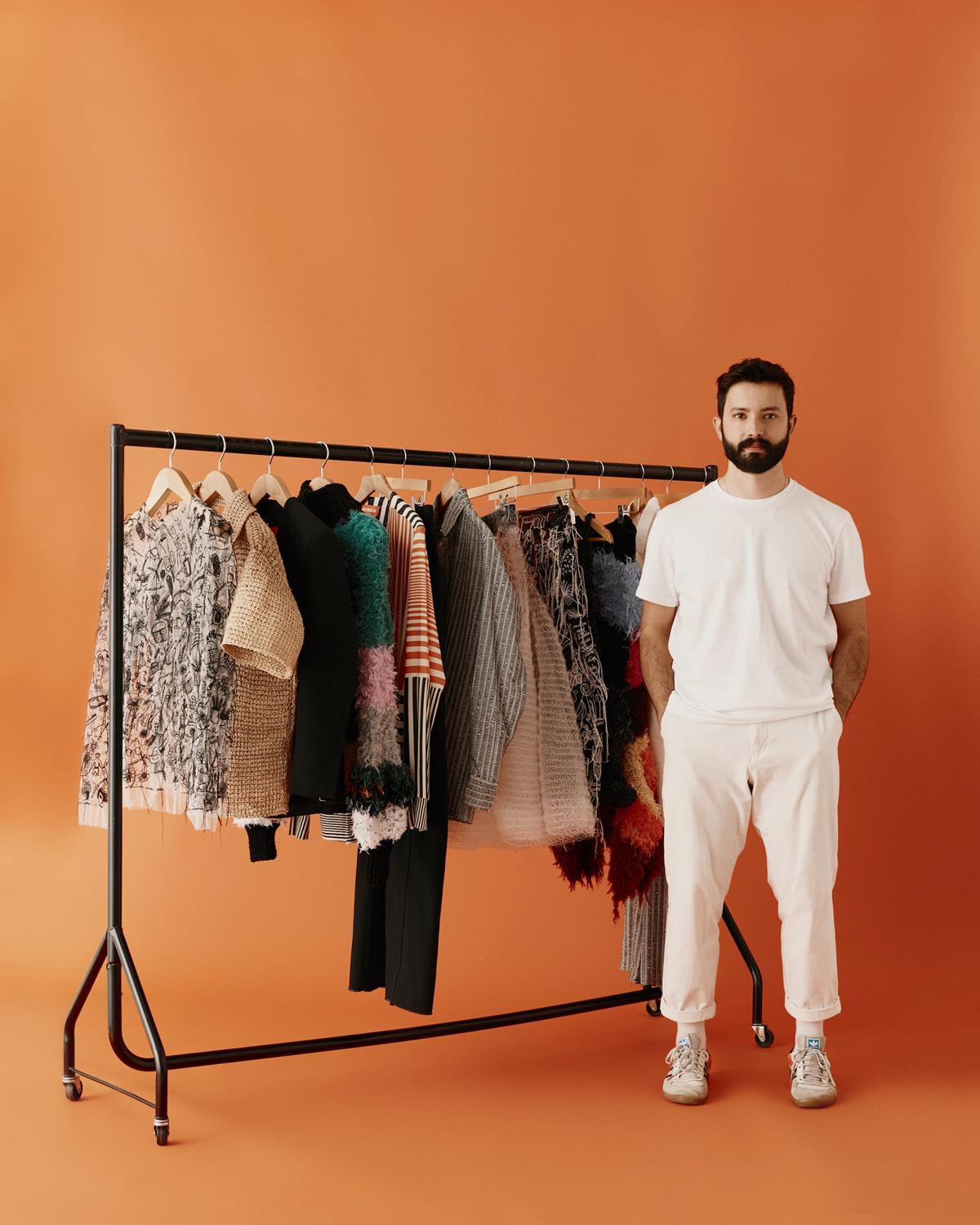 O estilista João Maraschin posa para foto em um fundo laranja cinza. Ele é um homem branco e jovem, de cabelo e barba preta, e usa uma camisa e uma calça branca. Está ao lado de uma arara com roupas da sua marca. - Metrópoles