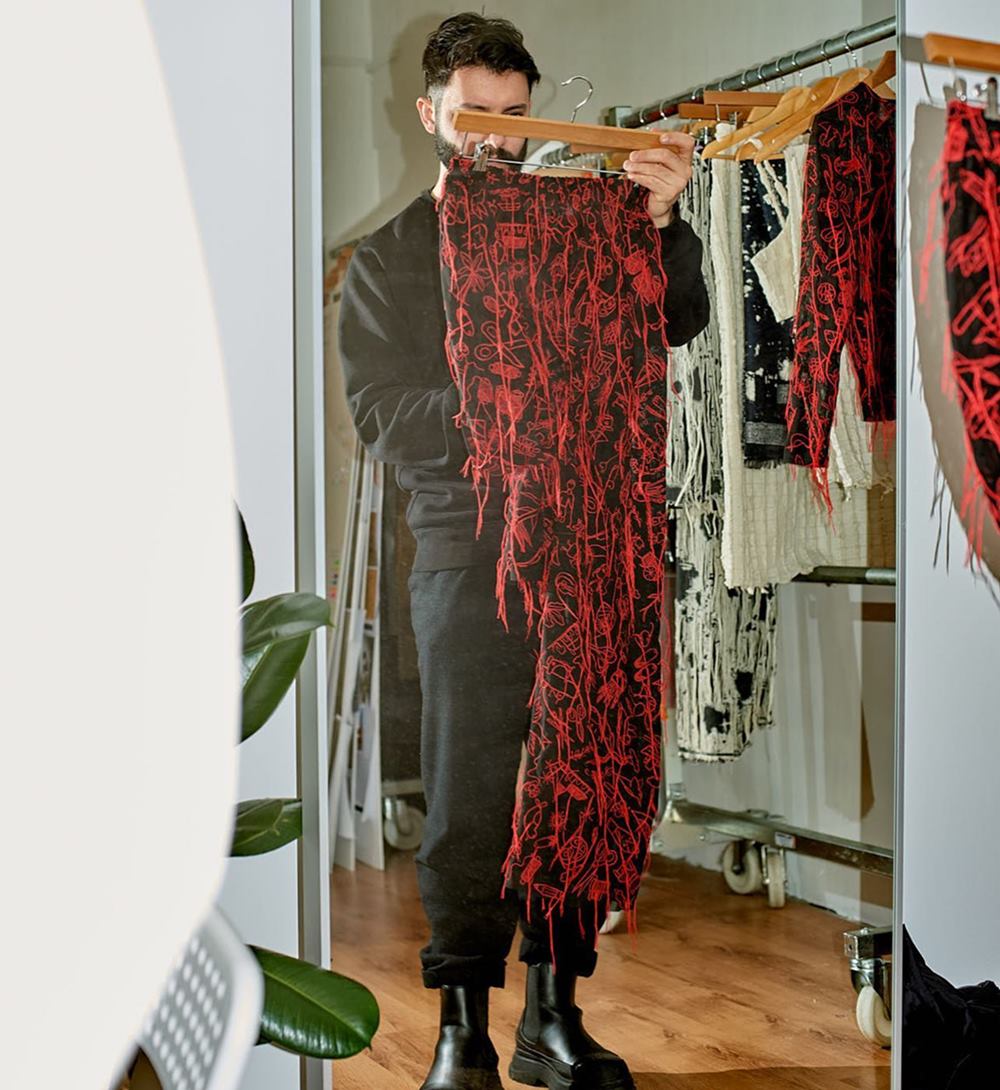 O estilista João Maraschin posa para foto segurando uma roupa feita por ele. Ele é um homem branco e jovem, de cabelo e barba preta, e usa uma camisa e uma calça preta. Está ao lado de uma arara com roupas da sua marca. - Metrópoles