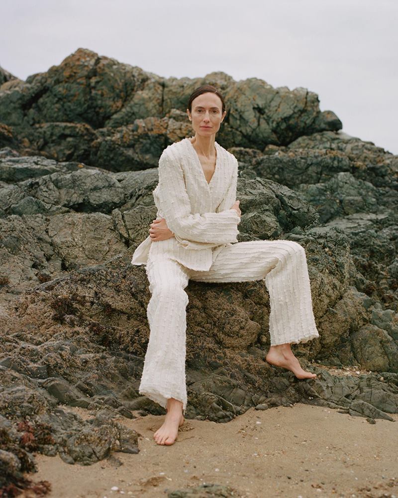 Fotos de campanha da marca do estilista João Maraschin. Na imagem, uma modelo branca e de meia idade, de cabelo liso loiro, está sentada nas pedras de uma praia. Ela usa um conjunto de crochê bee[ge composto por blusa, e calça. - Metrópoles