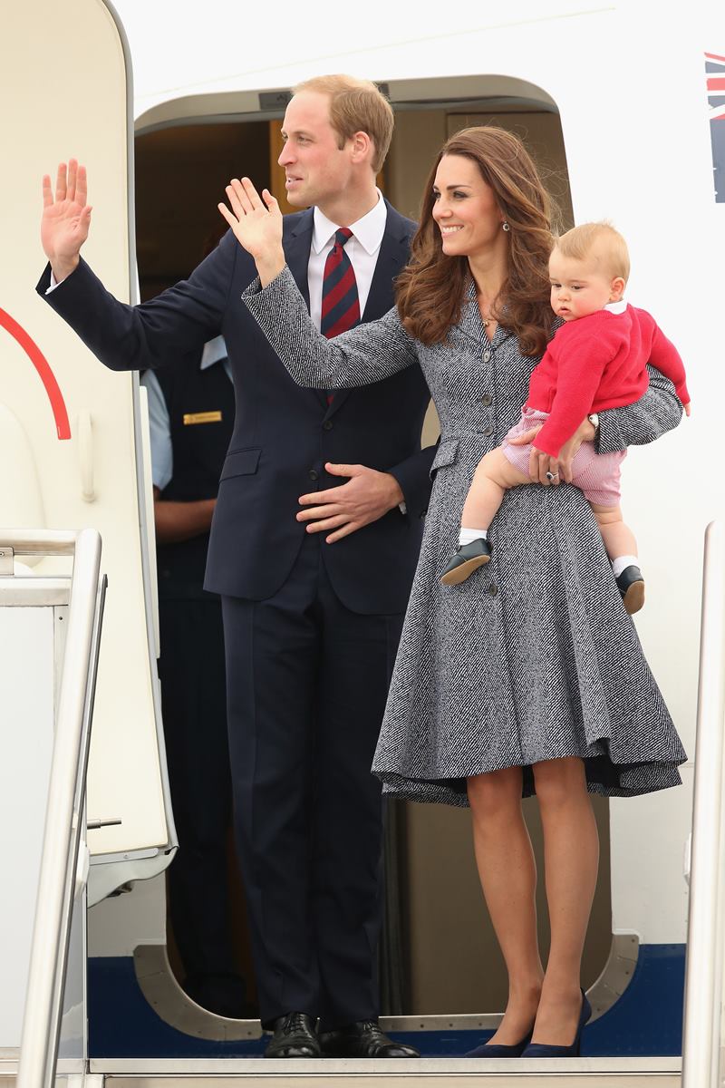 Foto colorida de homem branco e de terno, ao lado de mulher branca, com vestido cinza, segurando um bebê loiro com casaco vermelho. O casal dá tchau e está em frente a uma porta de avião - Metrópoles