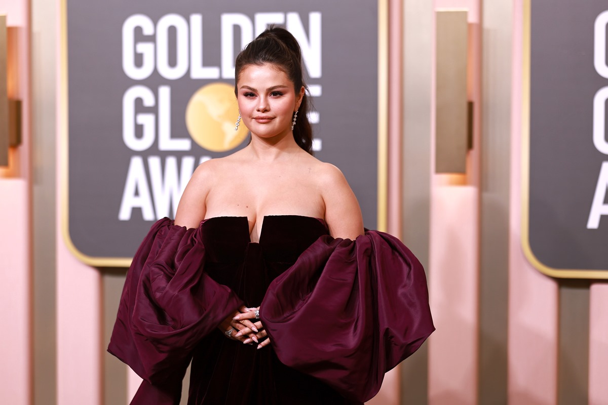 A cantora Selena Gomez no tapete vermelho do Globo de Ouro 2023. Ela é uma mulher branca e jovem, de cabelo ondulado preto, e usa um vestido preto de veludo com mangas roxas. - Metrópoles