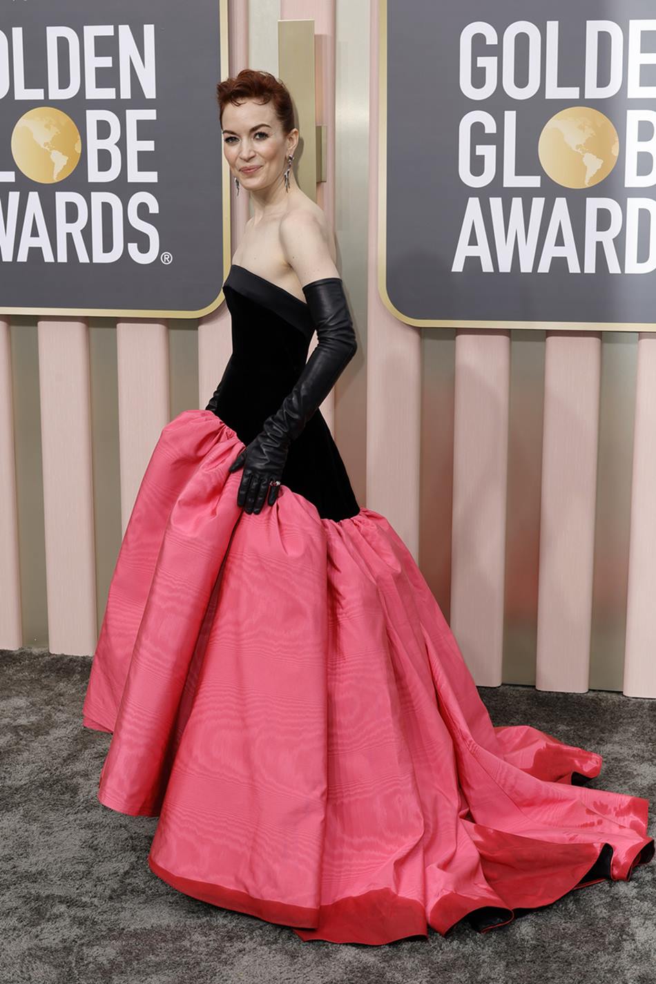 A atriz Britt Lower, um mulher branca e jovem, de cabelo liso ruivo, posa para foto no tapete vermelho do Globo de Ouro de 2023. Ela usa um vestido preto com uma saia bufante cor de rosa e luvas pretas de latex. - Metrópoles