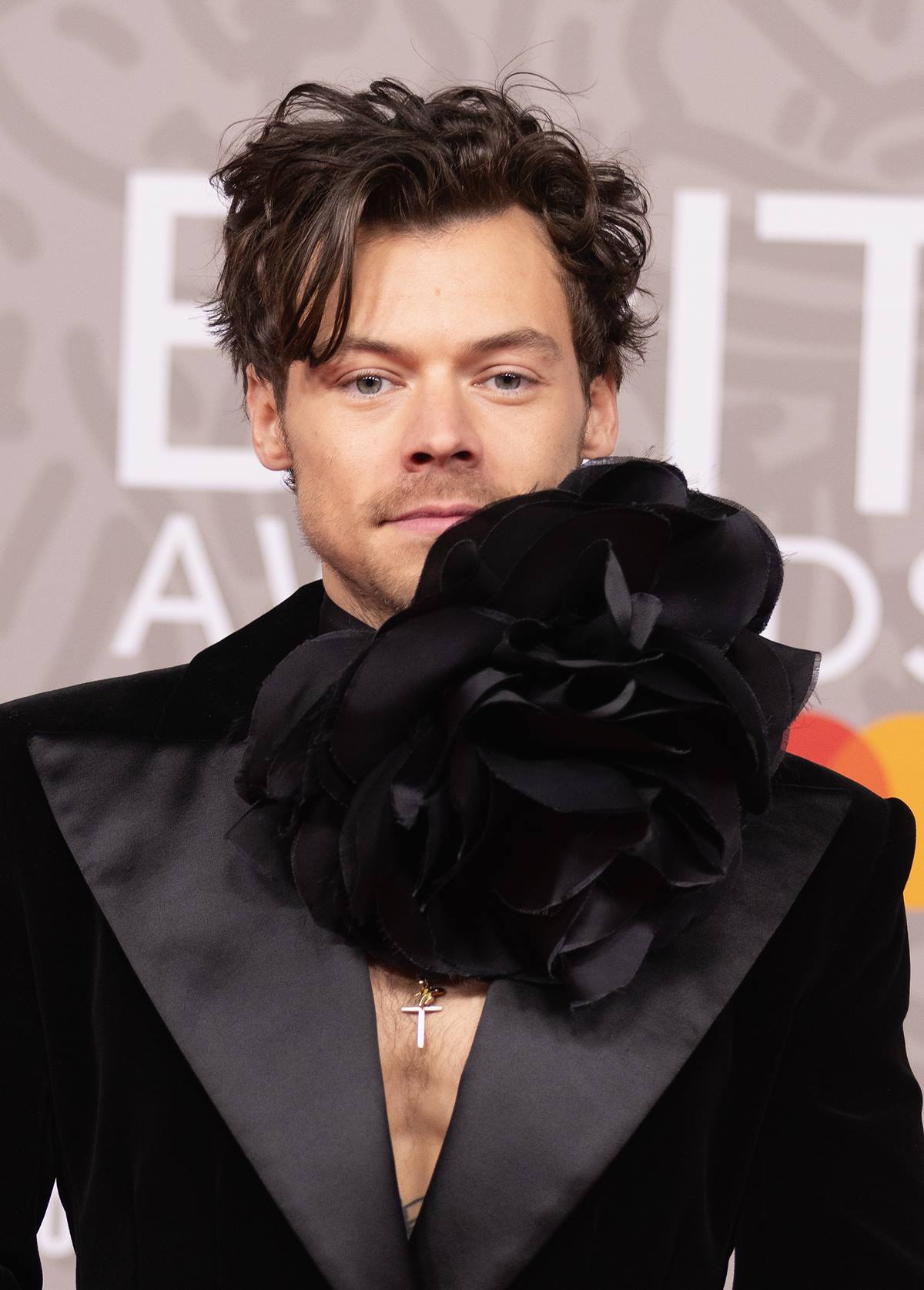 O cantor Harry Styles no tapete vermelho do BRIT Awards de 2023. Ele é um homem branco e jovem, de cabelo preto liso, e usa um blazer preto de veludo e uma gargantilha preta em formato de flor. - Metrópoles