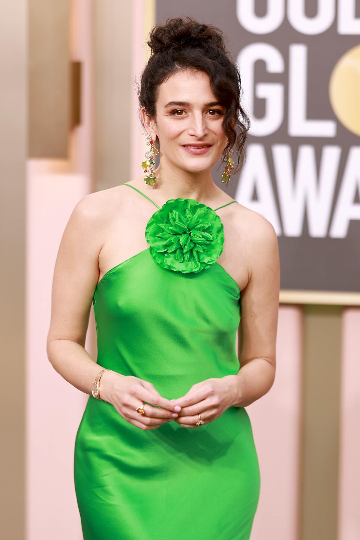 A atriz Jenny Slate, uma mulher branca e jovem, de cabelo cacheado curto, no tapete vermelho do Globo de Ouro de 2023. Ela usa um vestido de cetim verde com uma flor costurada perto do pescoço. A peça é da marca Rodarte. - Metrópoles